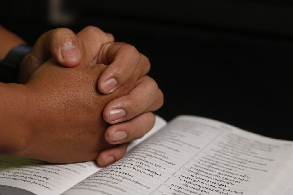 Close-up of hands in prayer over an open Khmer Bible, symbolizing faith and worship.