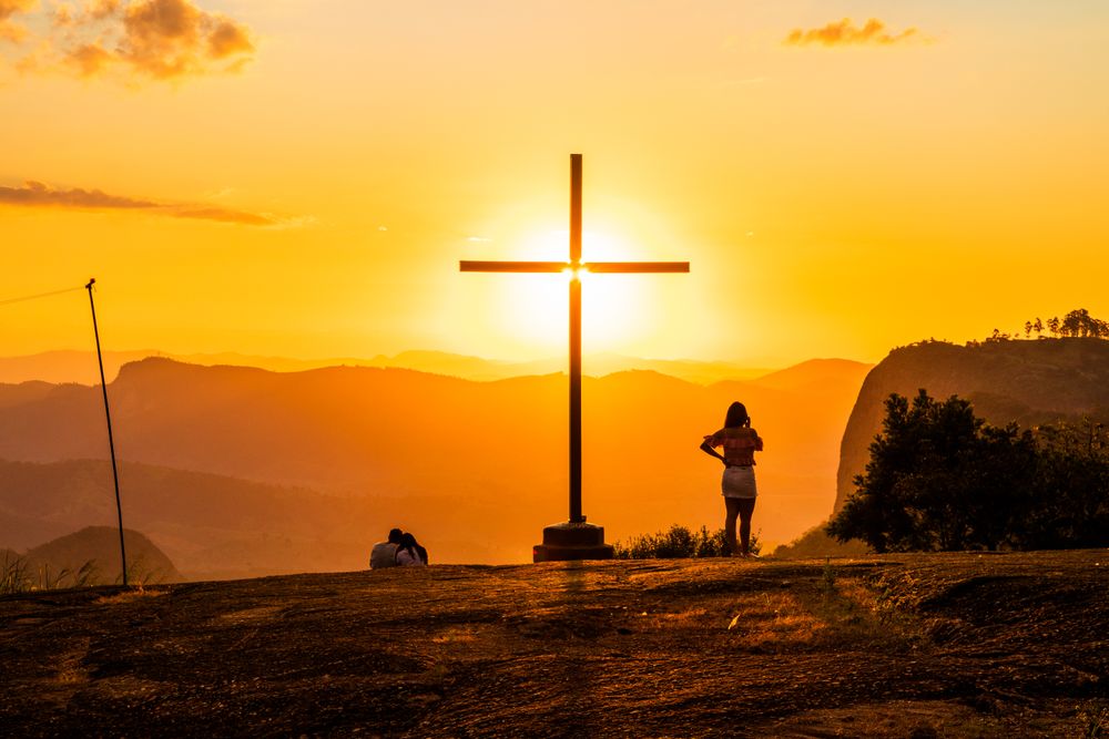 Back view of anonymous travelers standing near cross behind mountains under colorful sky with shining sun at sunset