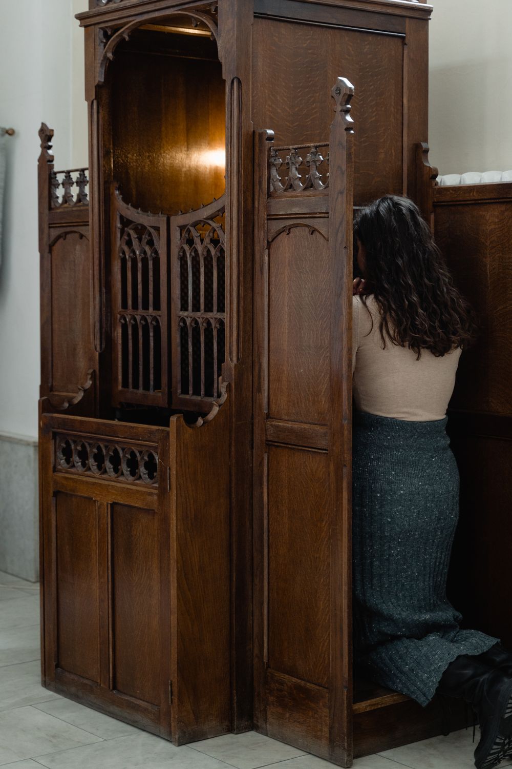 A woman kneels in a traditional wooden confessional booth, captured in an atmospheric church setting.