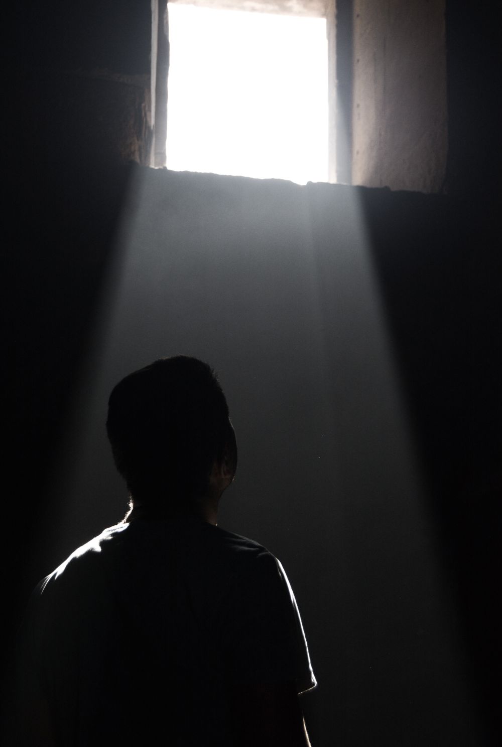 A man silhouetted in darkness looking towards a beam of light streaming through a window.
