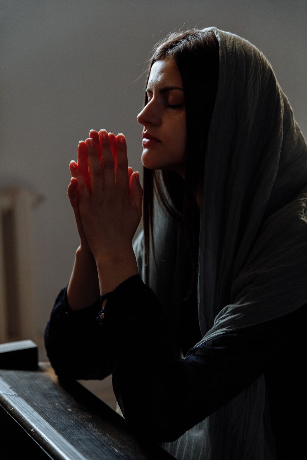 A woman with a headscarf prays solemnly inside a church, hands clasped and eyes closed.