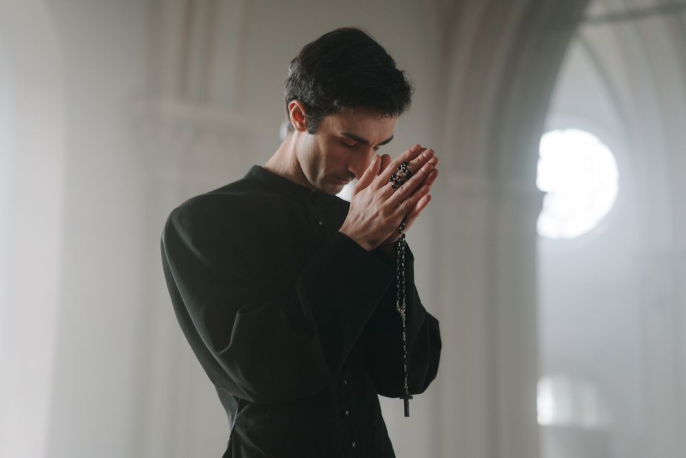 Devout man in black gown praying with a rosary inside a serene church setting.